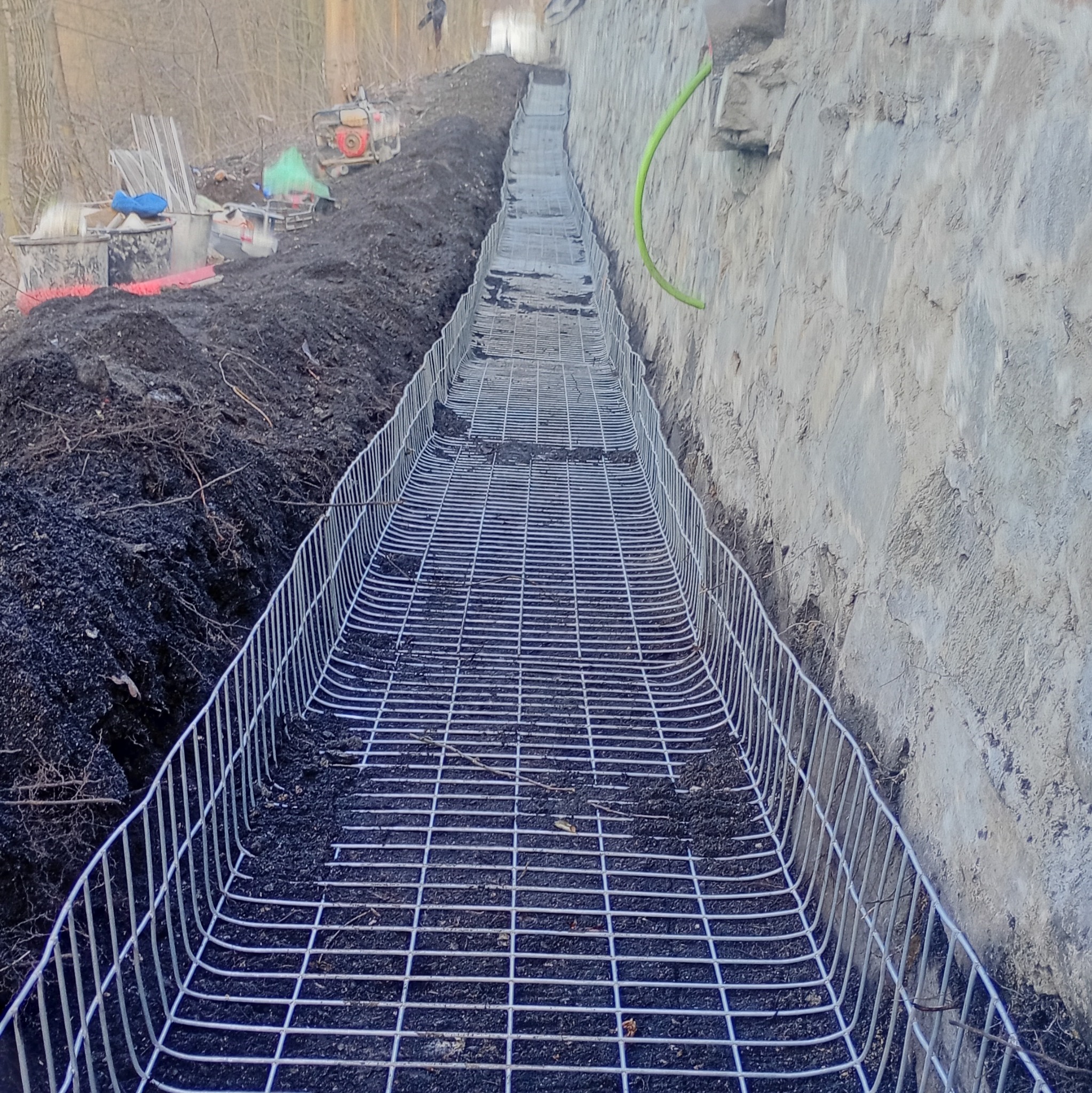 Przygotowanie terenu: metalowa siatka w wykopie wzdłuż betonowej ściany, narzędzia i sprzęt budowlany w tle. Etap prac ziemnych przed wylaniem fundamentów.