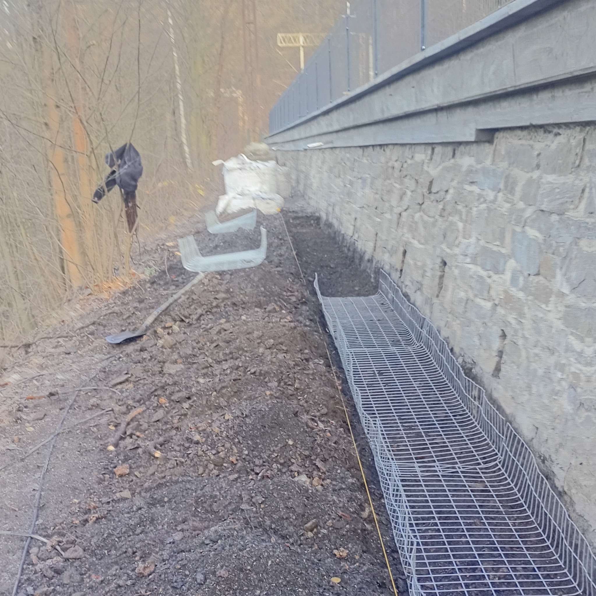 Przygotowanie terenu pod murek oporowy: wykop, siatka wzmacniająca, narzędzia. Prace ziemne w toku, widoczna ściana z kamienia i drzewa w tle.