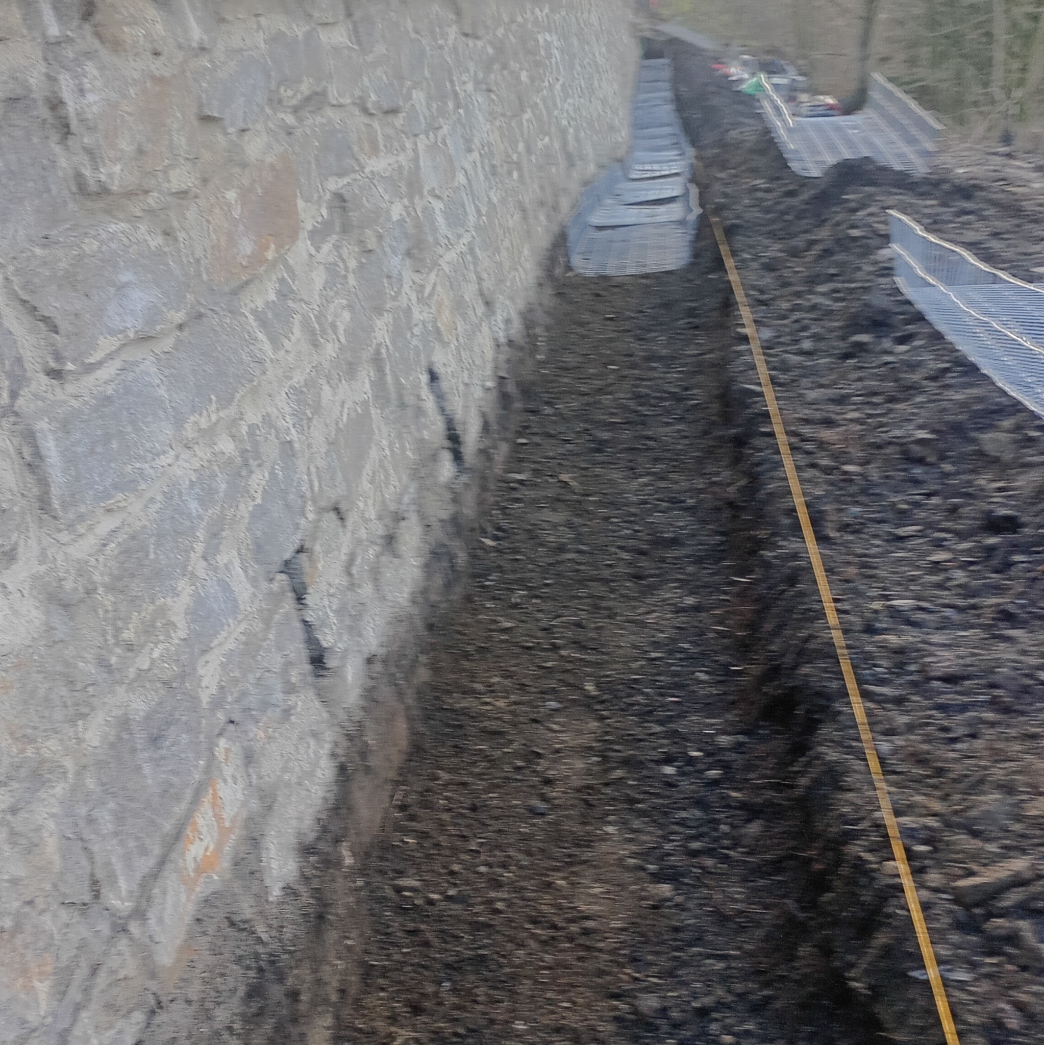 Wykopany rów wzdłuż kamiennego muru, zabezpieczony siatką. Widoczna żółta linia wyznaczająca kierunek. Prace ziemne w toku, przygotowanie pod instalacje.