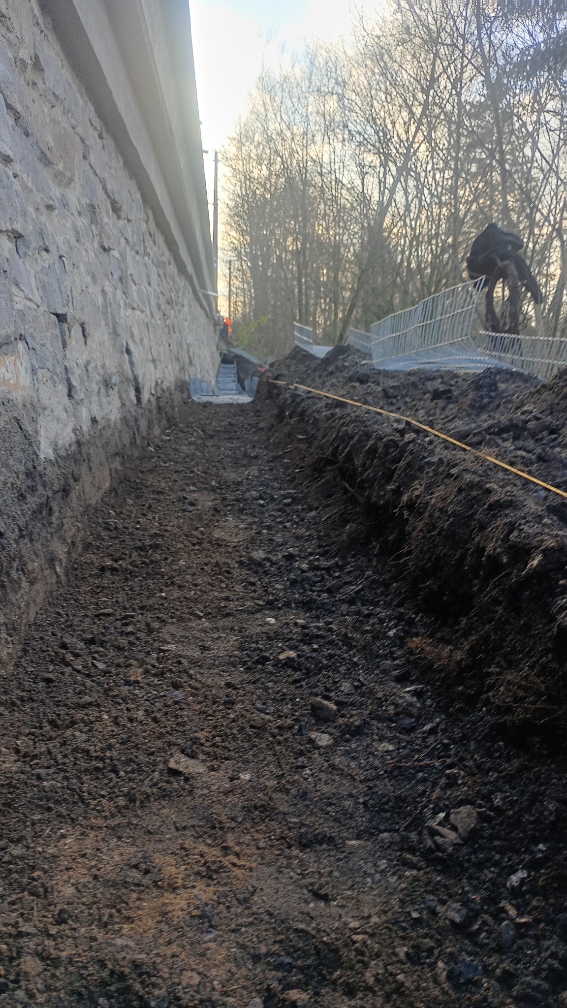 Wykopany rów wzdłuż kamiennego muru, widoczne zabezpieczenia wykopu i osoby pracujące w oddali. Przygotowanie terenu pod budowę lub instalacje.