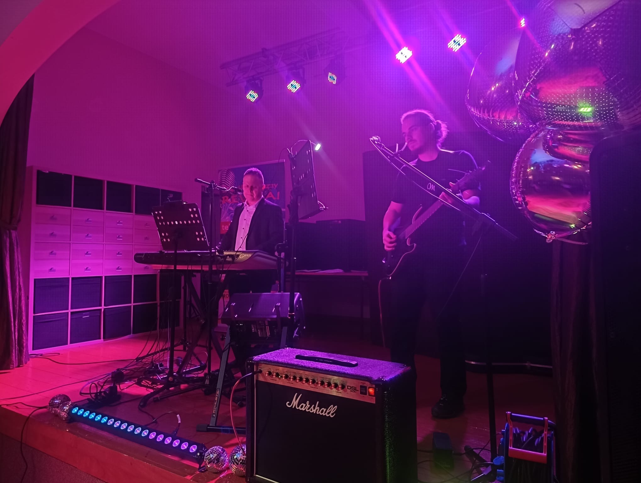 Zespół muzyczny na scenie: klawiszowiec w garniturze i gitarzysta z keytarem, oświetlenie sceniczne, wzmacniacz Marshall, dekoracje z kul dyskotekowych.