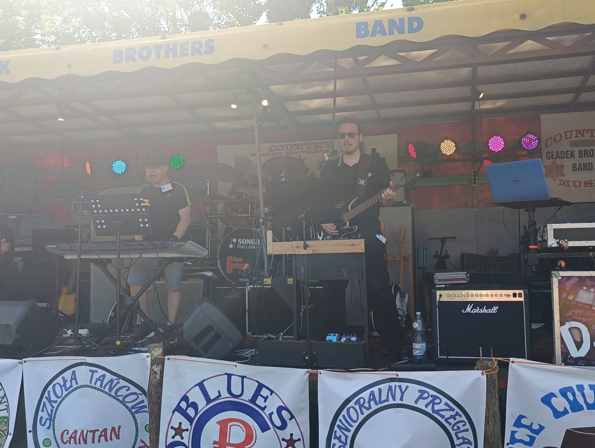 Zespół muzyczny na scenie plenerowej: gitarzysta w okularach słonecznych, klawiszowiec, perkusja Sonor, wzmacniacz Marshall, banery szkół tańca.