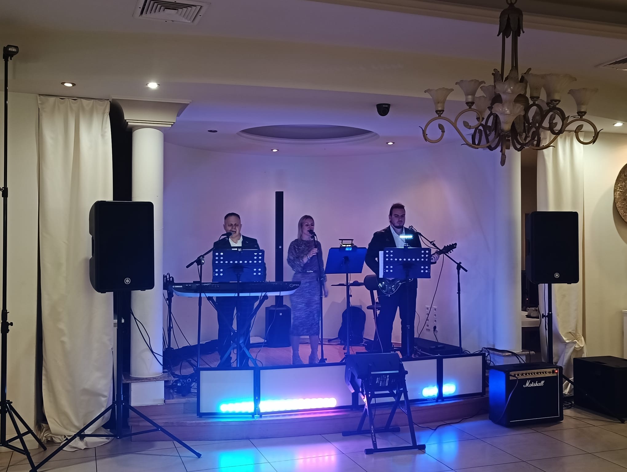 Zespół muzyczny na scenie z oświetleniem LED, grający na żywo. Wokalistka w sukience, klawiszowiec i gitarzysta. Kolumny głośnikowe Yamaha po bokach sceny.