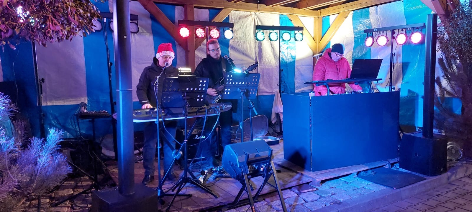 Zespół muzyczny gra na zewnątrz pod zadaszeniem, oświetlony kolorowymi światłami. Jeden z muzyków ma czapkę Mikołaja. Widoczny keyboard, gitara i stanowisko DJ.