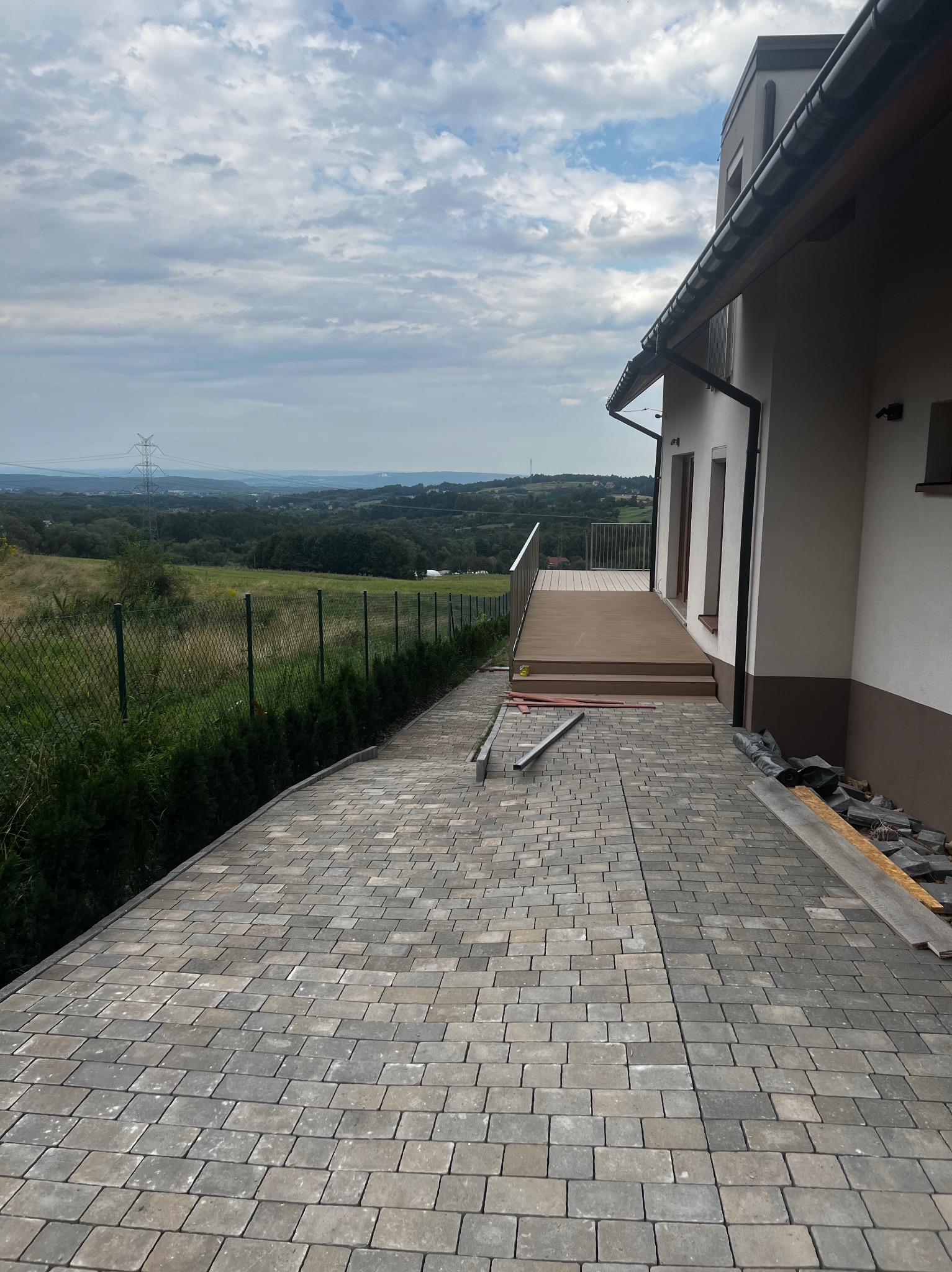 Ułożona kostka brukowa na podjeździe domu z widokiem na krajobraz. Częściowo widoczny taras i elewacja budynku. Prace brukarskie w toku, widoczne narzędzia i materiały.