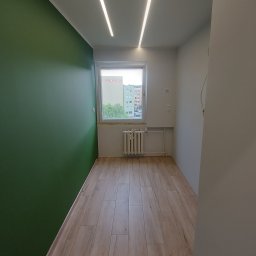 DJK usługi remontowo-budowlane Krzysztof Jacek Dąbrowa - Pokój po remoncie: zielona ściana, drewnopodobna podłoga, okno z widokiem na budynki, grzejnik, nowoczesne oświetlenie sufitowe. Minimalistyczny design.