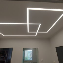 DJK usługi remontowo-budowlane Krzysztof Jacek Dąbrowa - Nowoczesne oświetlenie sufitowe LED w kształcie kwadratów, tworzące geometryczny wzór. Minimalistyczny design w jasnym wnętrzu z widocznymi drzwiami i oknem.