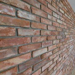 DJK usługi remontowo-budowlane Krzysztof Jacek Dąbrowa - Ściana z czerwonej cegły klinkierowej, ułożonej z przesunięciem, z widoczną szarą fugą. Cegły o zróżnicowanej kolorystyce, od jasnych do ciemnych odcieni czerwieni. Widok z boku, perspektywa.