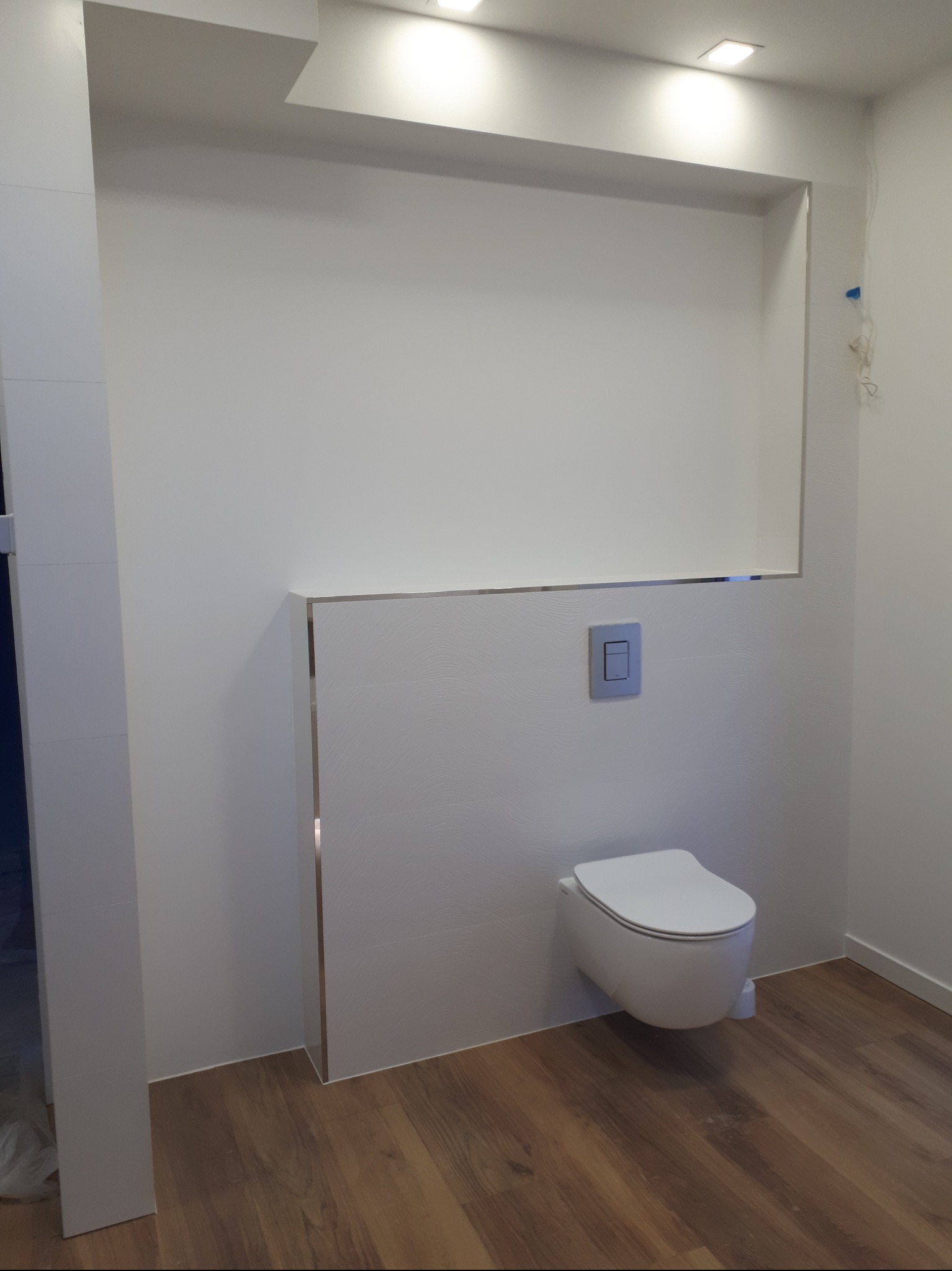 Nowoczesna, podwieszana toaleta w wykończonej łazience z drewnopodobną podłogą i białymi płytkami o delikatnej strukturze. Minimalistyczny design, oświetlenie punktowe.