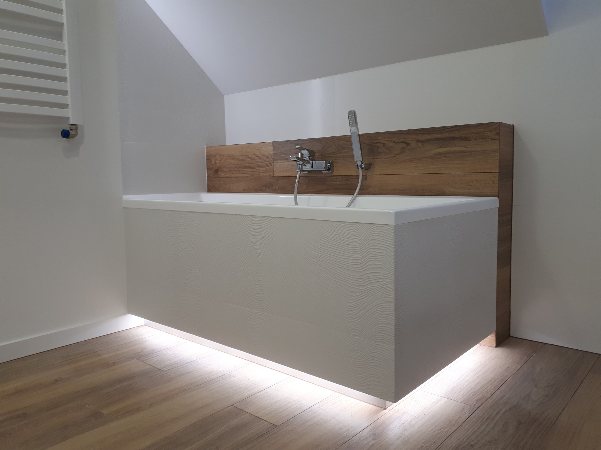 Nowoczesna łazienka na poddaszu: biała wanna z podświetleniem LED, drewniane panele za baterią, grzejnik drabinkowy i drewnopodobna podłoga.