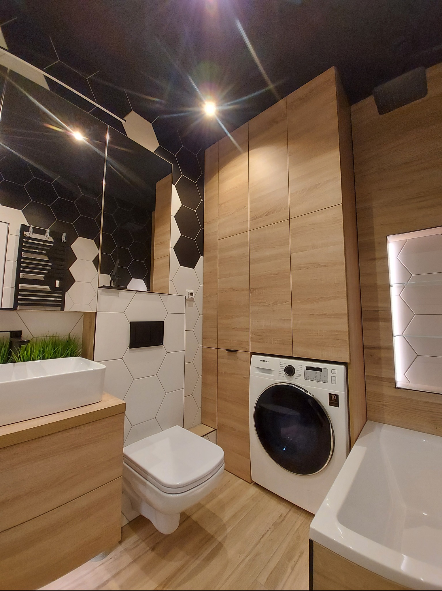 Nowoczesna łazienka z heksagonalnymi płytkami, drewnianymi szafkami, pralką Samsung i białym wyposażeniem. Ciemny sufit i podłoga imitująca drewno.