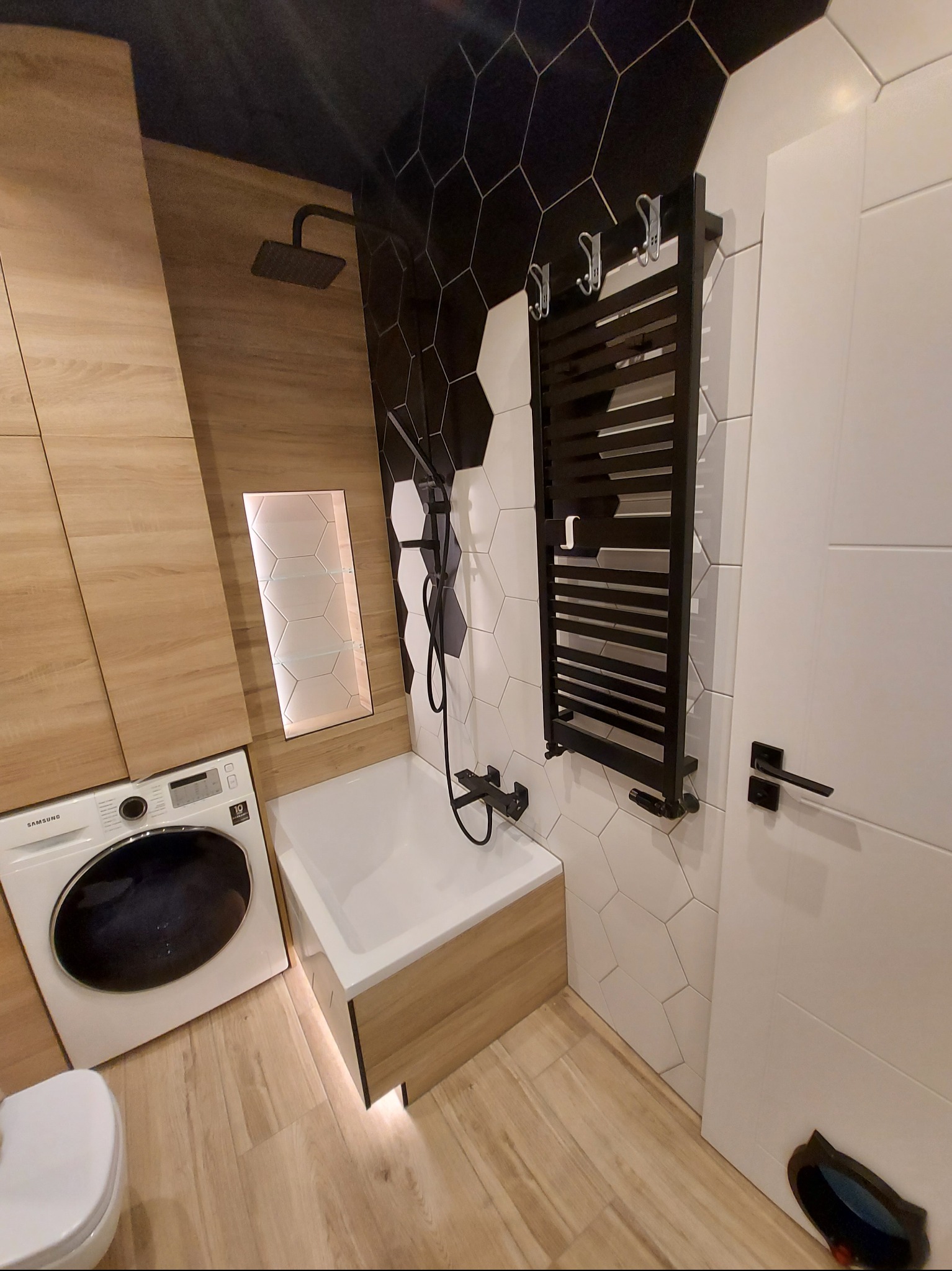 Nowoczesna łazienka z heksagonalnymi płytkami, drewnianymi akcentami i czarną armaturą. Widoczna pralka, wanna i grzejnik drabinkowy. Całość utrzymana w jasnej kolorystyce.
