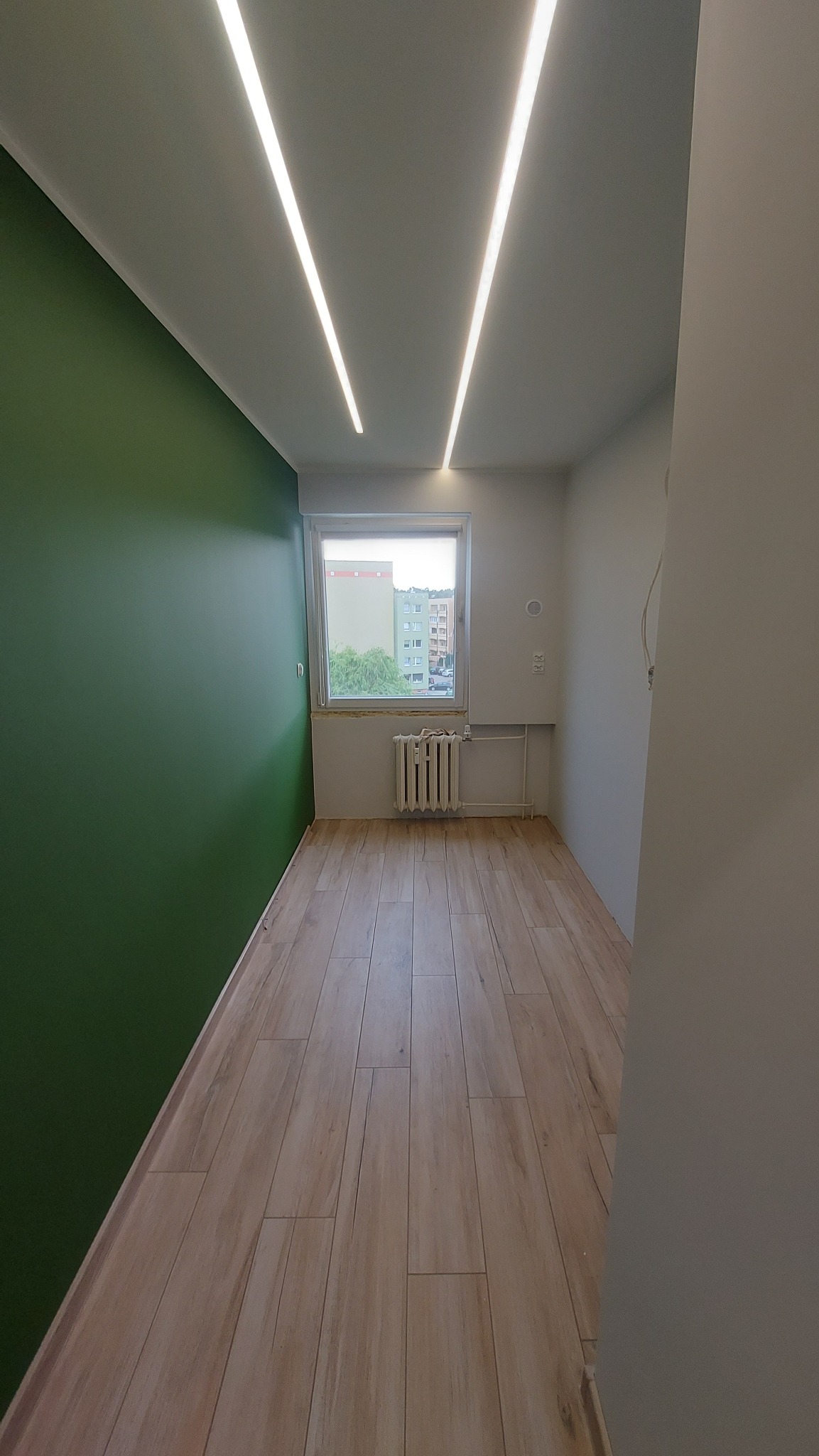 Pokój po remoncie: zielona ściana, drewnopodobna podłoga, okno z widokiem na budynki, grzejnik, nowoczesne oświetlenie sufitowe. Minimalistyczny design.