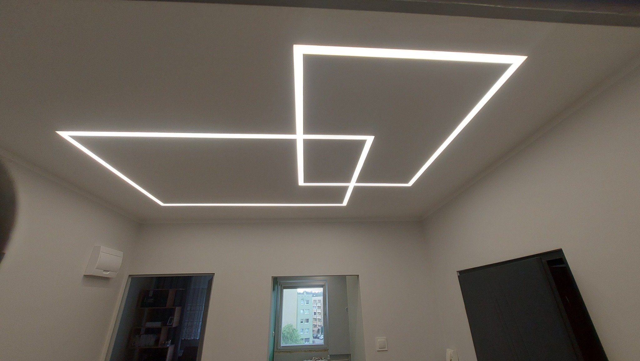 Nowoczesne oświetlenie sufitowe LED w kształcie kwadratów, tworzące geometryczny wzór. Minimalistyczny design w jasnym wnętrzu z widocznymi drzwiami i oknem.