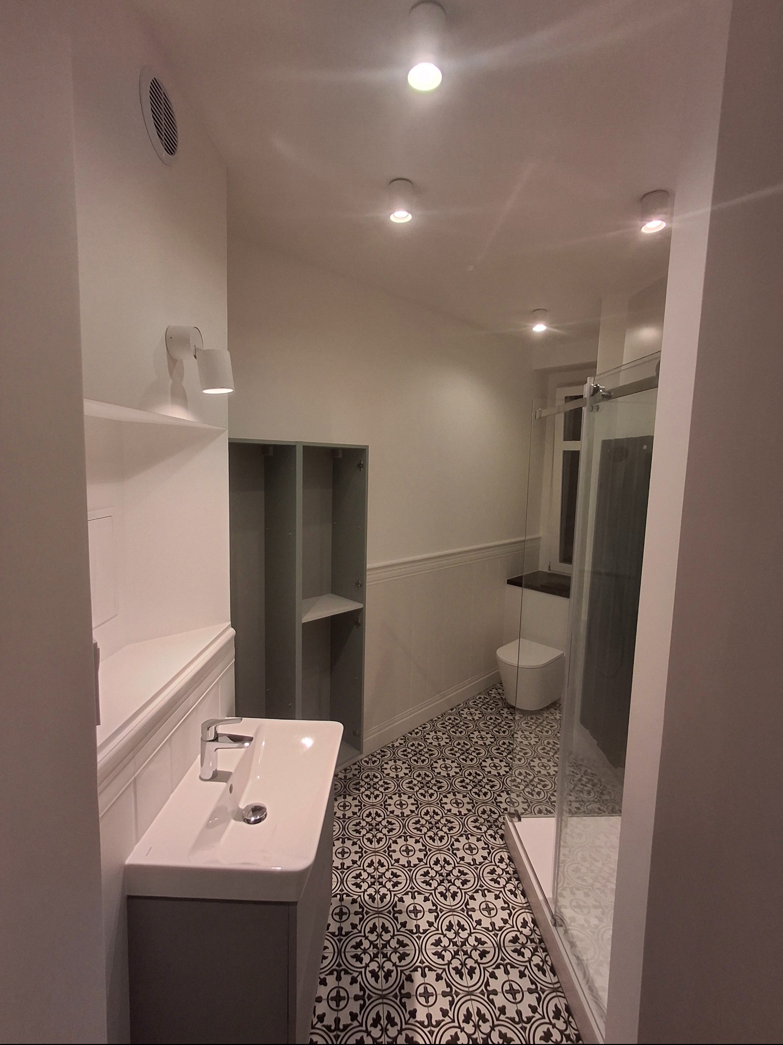 Wykończona łazienka w bieli i szarości z wzorzystą podłogą. Widoczna umywalka z szafką, kabina prysznicowa i toaleta. Oświetlenie punktowe.