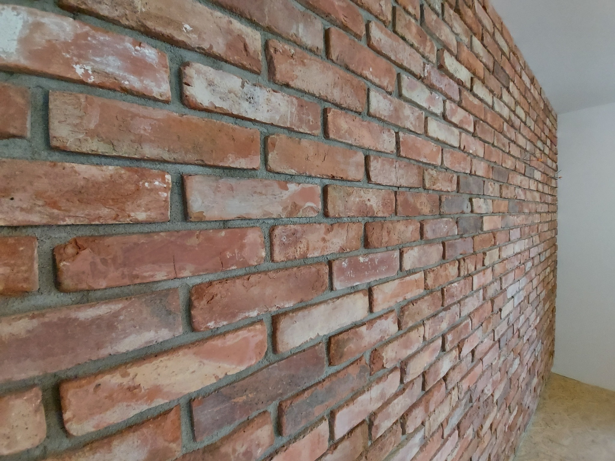 Ściana z czerwonej cegły klinkierowej, ułożonej z przesunięciem, z widoczną szarą fugą. Cegły o zróżnicowanej kolorystyce, od jasnych do ciemnych odcieni czerwieni. Widok z boku, perspektywa.