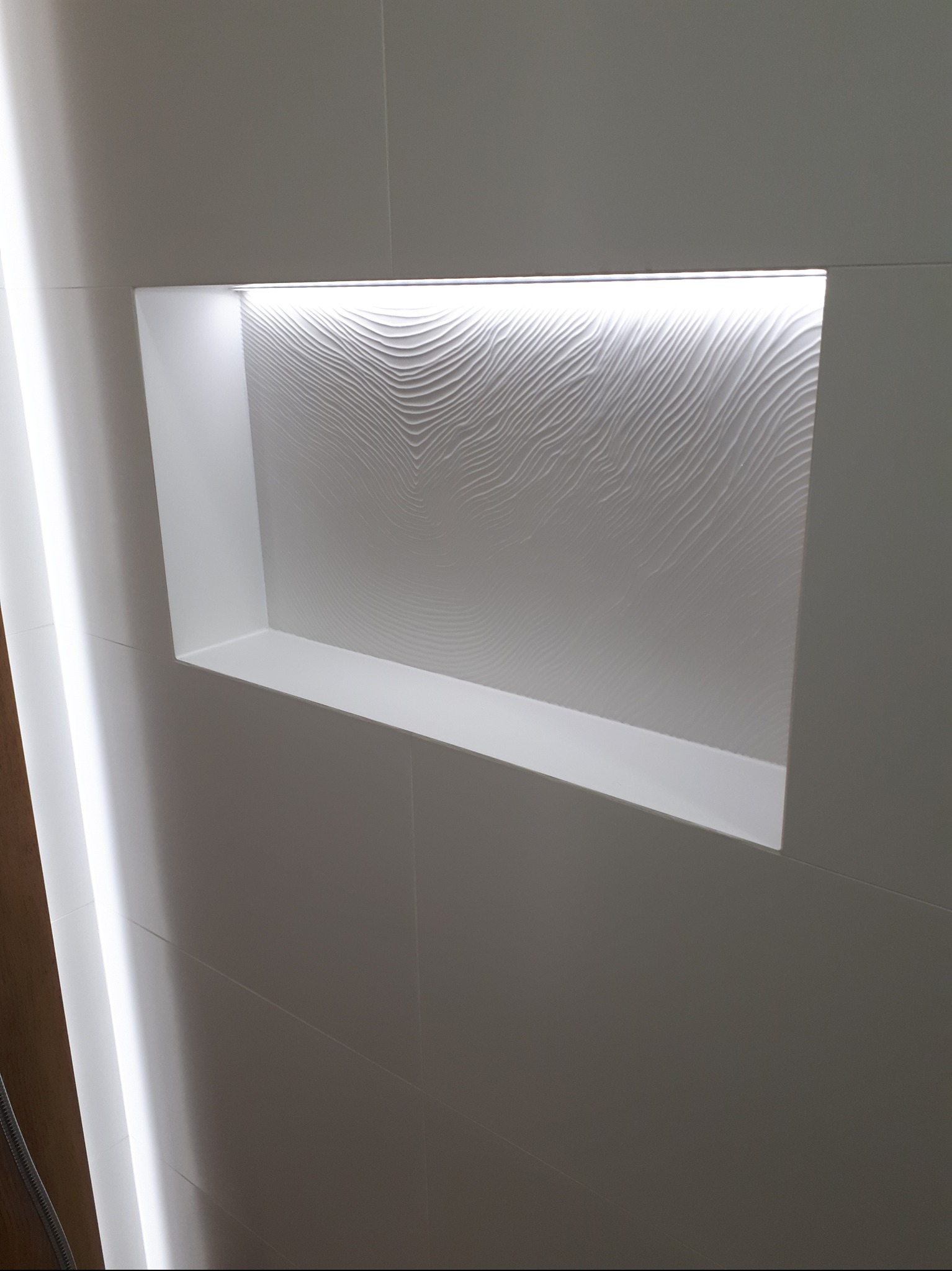Biała, podświetlana wnęka z dekoracyjną fakturą na ścianie wykończonej płytkami w łazience. Minimalistyczny design z oświetleniem LED.