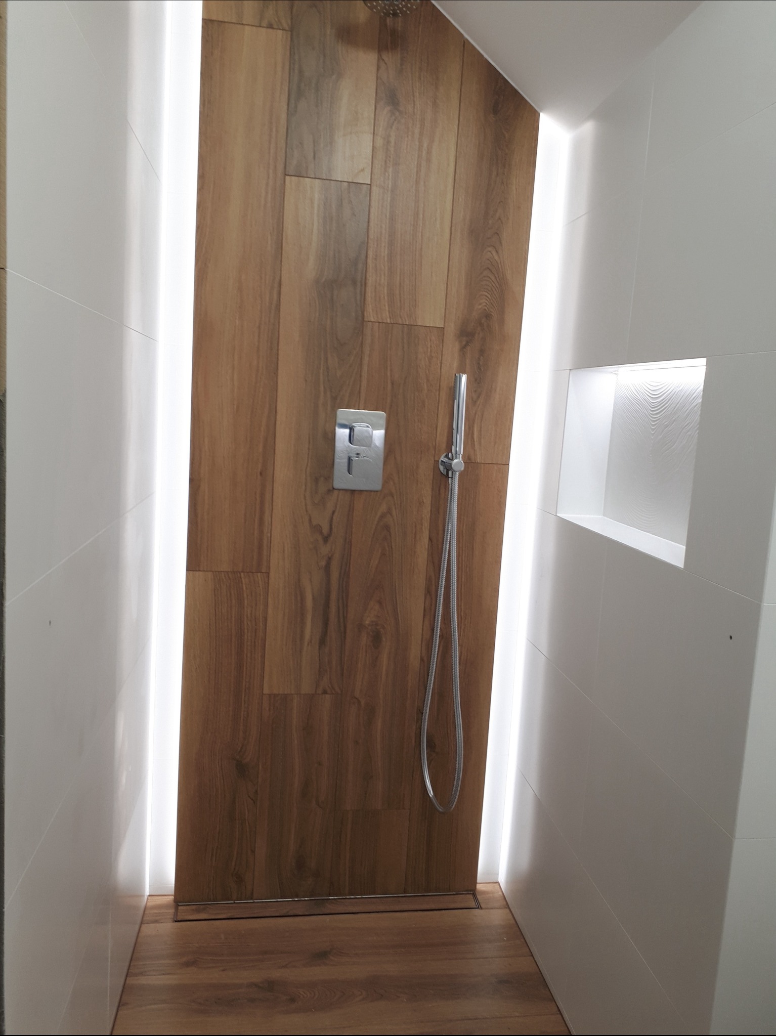 Nowoczesna kabina prysznicowa z drewnopodobnymi płytkami, białymi ścianami i subtelnym oświetleniem LED. Widoczna armatura i wnęka w ścianie.