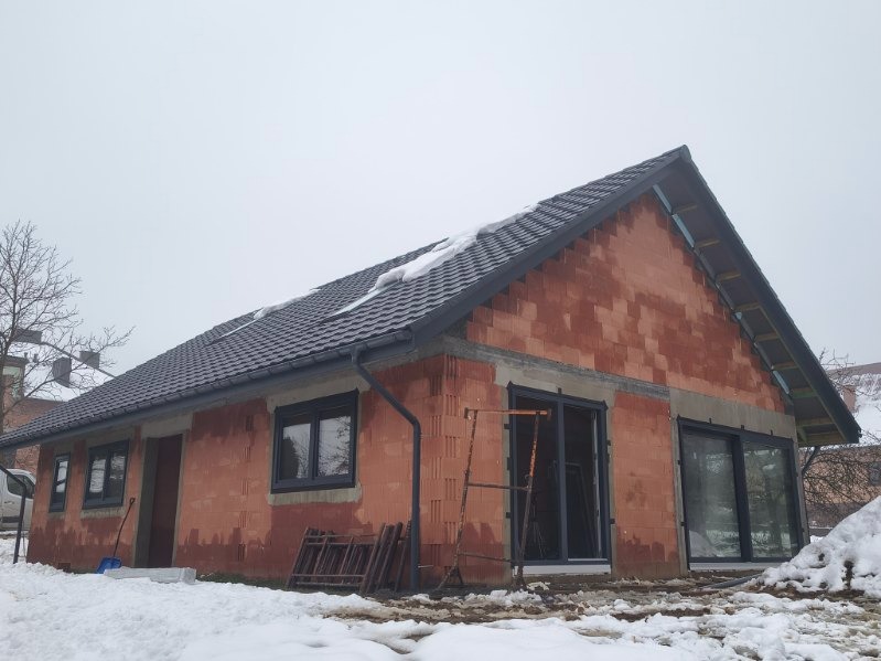 Dom w stanie surowym zamkniętym, z czarną dachówką i szarą stolarką okienną. Widoczne rusztowanie i śnieg. Ujęcie z zewnątrz, w pochmurny dzień.