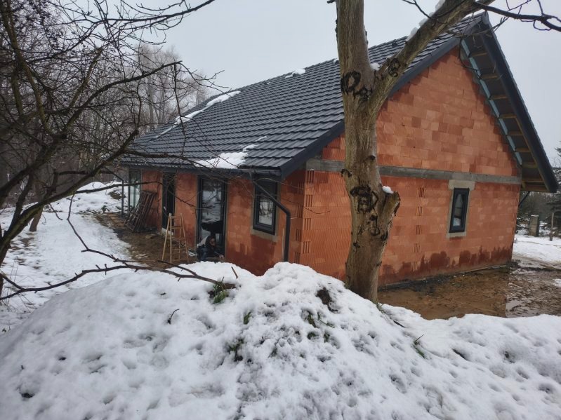 Surowy stan domu z czerwonej cegły w zimowej scenerii. Widoczne czarne okna, fragment dachu z dachówką i drzewo na pierwszym planie. Etap budowy w trakcie zimy.