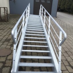 FHU Stalbud - Zewnętrzne, metalowe schody z srebrnymi poręczami prowadzące do szarego budynku z brązową bramą garażową. Widok z dołu, podkreślający solidną konstrukcję.
