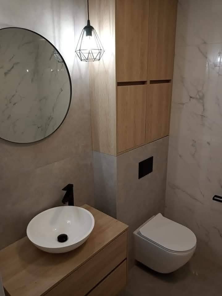 Nowoczesna łazienka z okrągłym lustrem, drewnianą szafką pod umywalką i wiszącą toaletą. Marmurowe płytki na ścianach, designerska lampa wisząca.