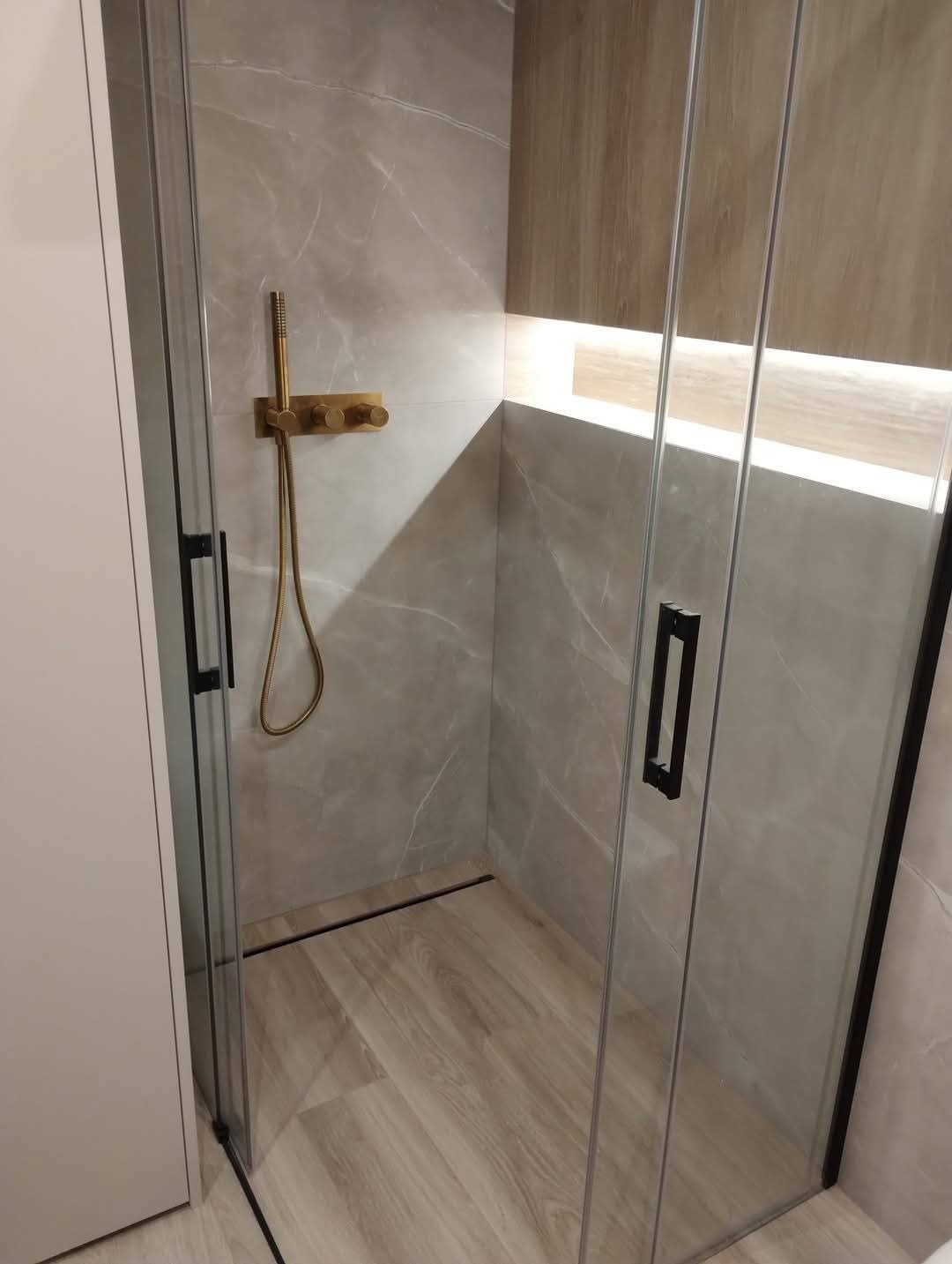 Nowoczesna kabina prysznicowa z szarymi płytkami imitującymi marmur, złotą armaturą i drewnopodobną zabudową. Oświetlenie LED podkreśla elegancję wykończenia.
