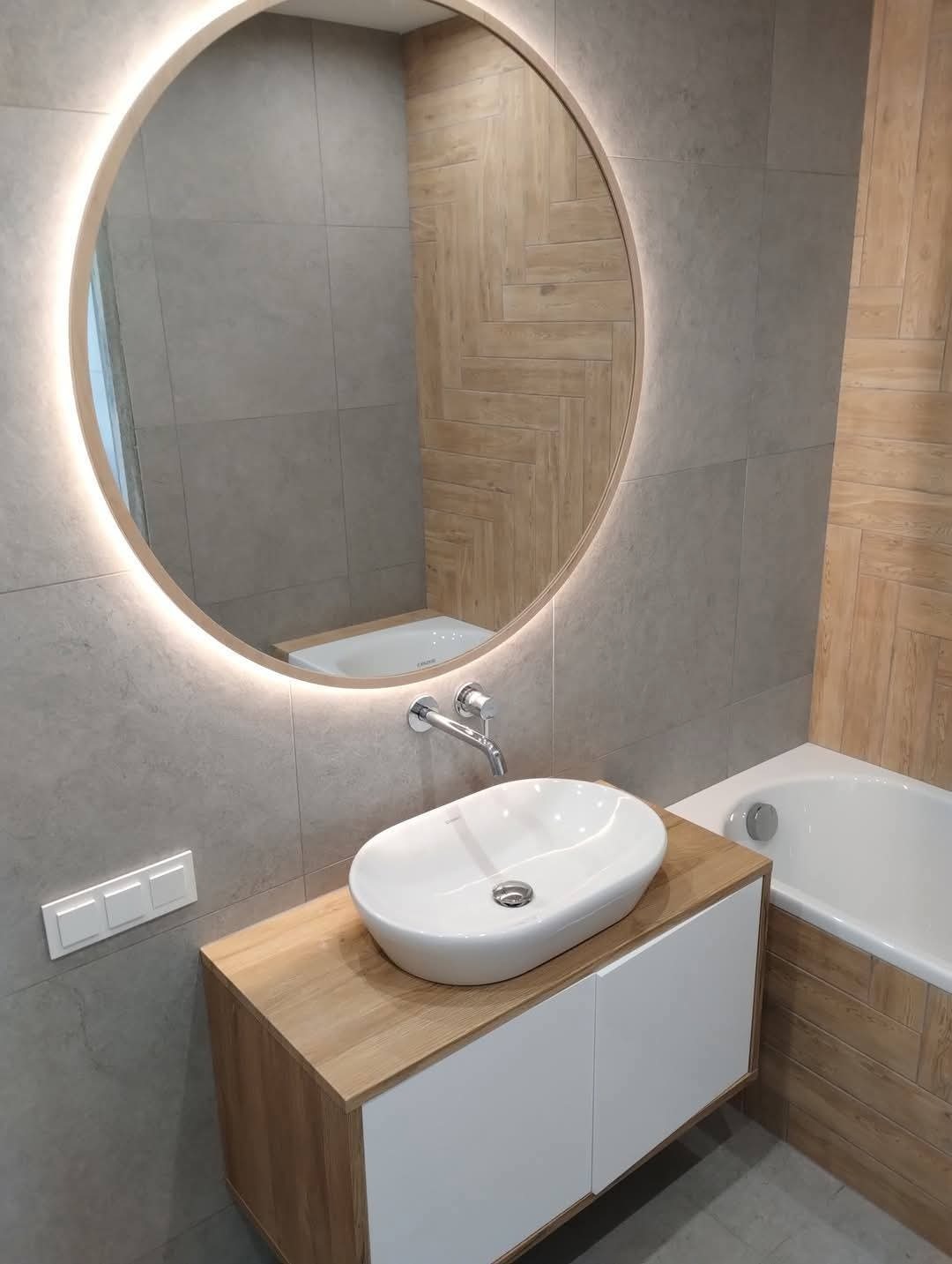 Nowoczesna łazienka z okrągłym lustrem LED, umywalką nablatową na szafce z drewnianym blatem i wanną obudowaną drewnopodobnymi płytkami.