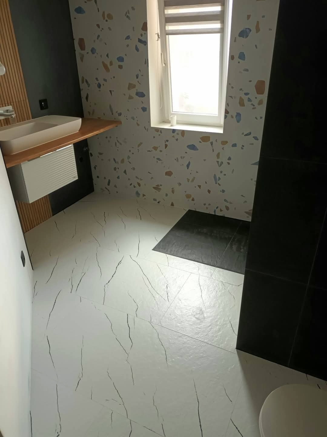 Nowoczesna łazienka po remoncie: białe płytki podłogowe z czarnymi żyłami, ściana z wzorem lastryko, drewniany blat z umywalką i czarne płytki w strefie prysznica.