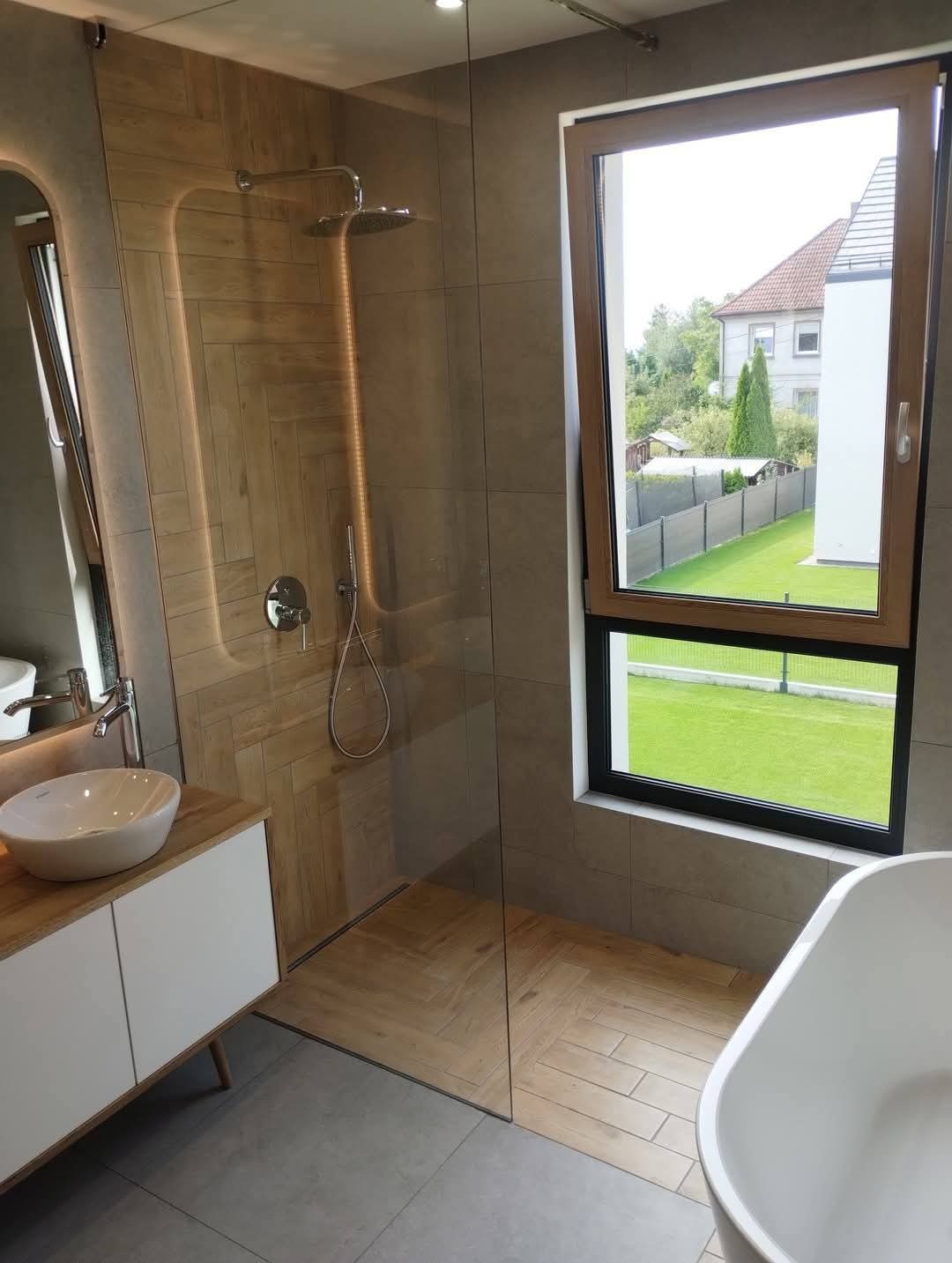 Nowoczesna łazienka z kabiną prysznicową typu walk-in, wykończona płytkami imitującymi drewno ułożonymi w jodełkę. Widok na zieleń przez okno.