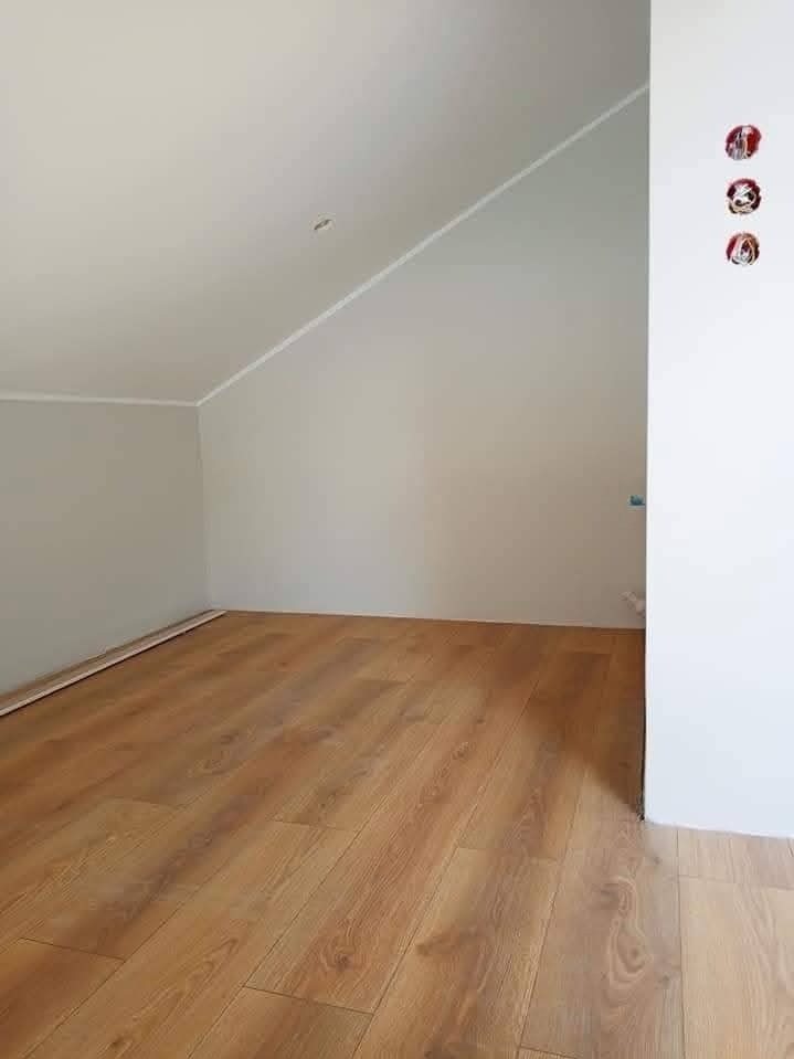 Poddasze po remoncie: świeżo ułożona, jasna podłoga z paneli, pomalowane ściany w odcieniach bieli i szarości. Widoczne gniazdka elektryczne i listwy przypodłogowe.