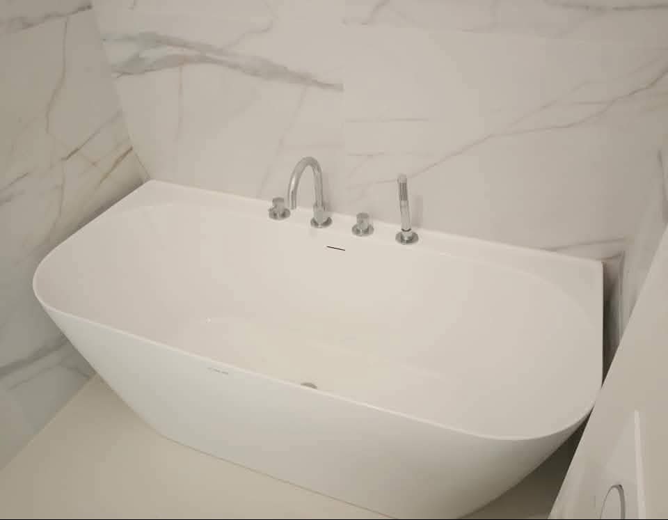 Biała, wolnostojąca wanna z chromowaną armaturą na tle ściany wyłożonej marmurowymi płytami. Minimalistyczny design, nowoczesna łazienka po remoncie.