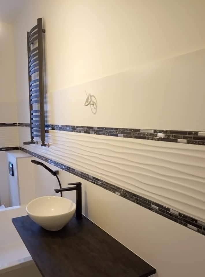 Nowoczesna łazienka z umywalką nablatową, czarną armaturą i grzejnikiem drabinkowym. Dekoracyjne, faliste płytki ścienne z mozaiką. Widoczne przewody elektryczne.