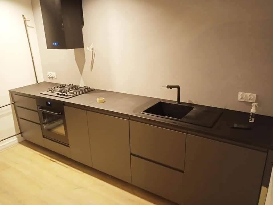 Nowoczesna, minimalistyczna kuchnia w odcieniach szarości z czarnymi akcentami: bateria, zlew, płyta. Jasna podłoga, gładkie fronty szafek, brak uchwytów.