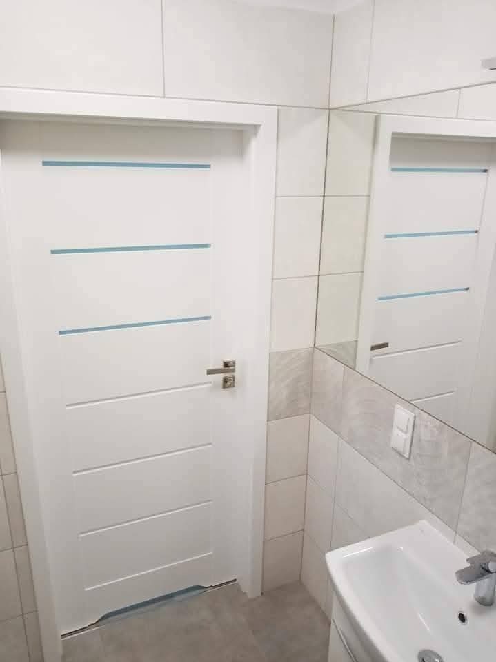 Białe drzwi z poziomymi, błękitnymi akcentami w łazience z jasnymi płytkami i lustrem, odbijającym drzwi. Widoczna umywalka i bateria.