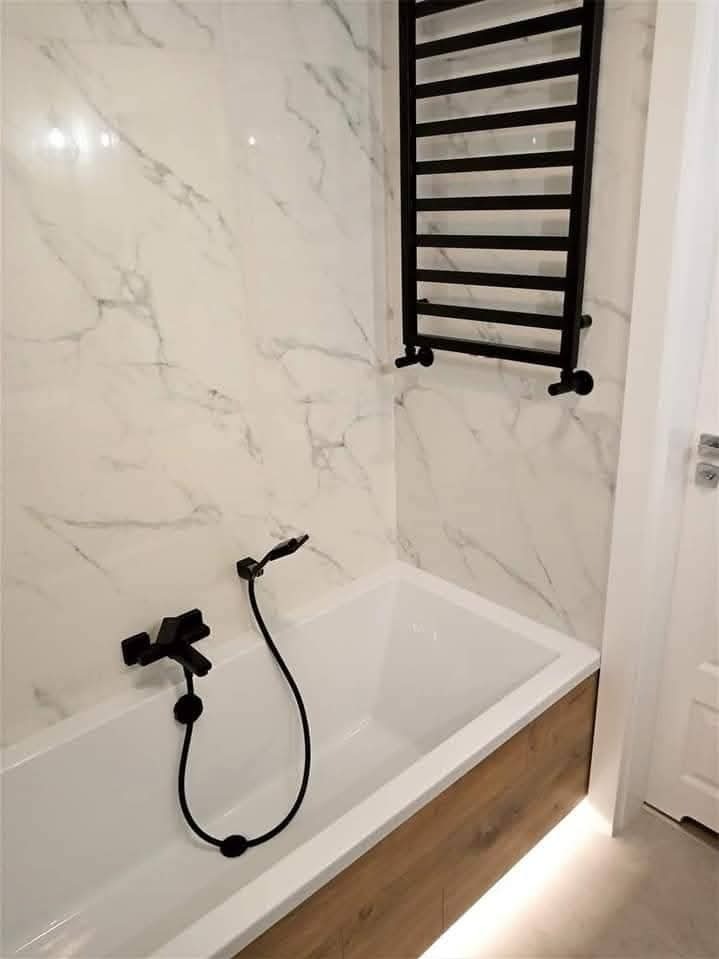 Biała wanna z drewnianym panelem i czarną armaturą na tle marmurowej ściany. Grzejnik drabinkowy w kolorze czarnym. Podświetlenie LED pod wanną.