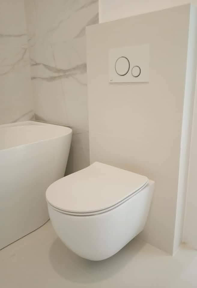 Nowoczesna, podwieszana toaleta w minimalistycznej łazience z marmurową ścianą i wolnostojącą wanną. Biała armatura i subtelne oświetlenie.