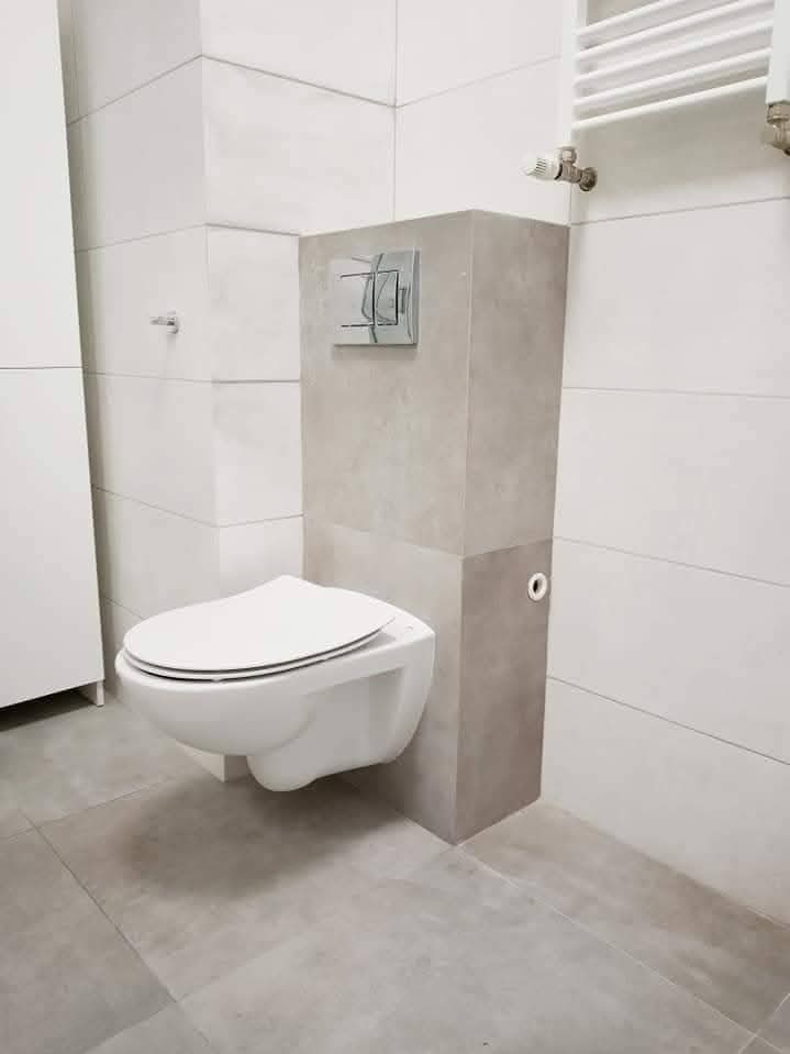 Nowoczesna toaleta podwieszana z szarą obudową, białe płytki na ścianach i podłodze, minimalistyczny design, chromowany przycisk spłukiwania, grzejnik drabinkowy w tle.