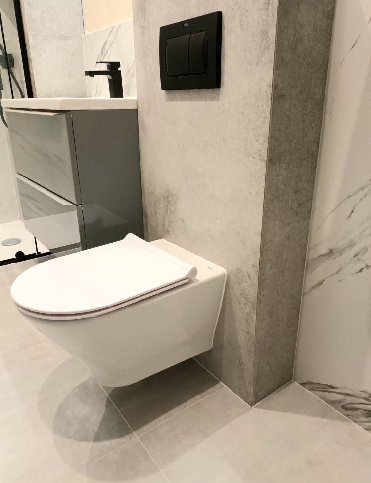 Nowoczesna toaleta podwieszana z czarnym przyciskiem spłukującym na tle szarej, marmurkowej ściany. Umywalka z szafką w tle. Minimalistyczny design.