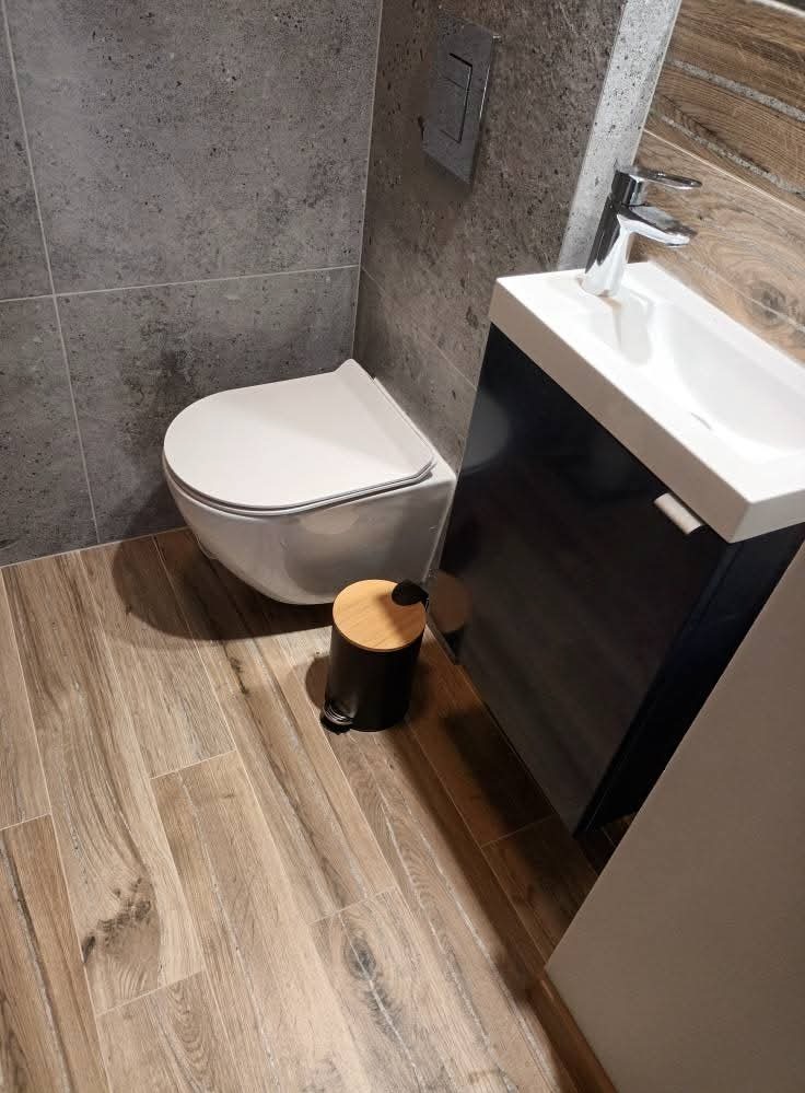Nowoczesna łazienka z szarą, teksturowaną ścianą, drewnopodobną podłogą, podwieszaną toaletą i szafką z umywalką. Uwagę zwraca kosz na śmieci z drewnianą pokrywką.
