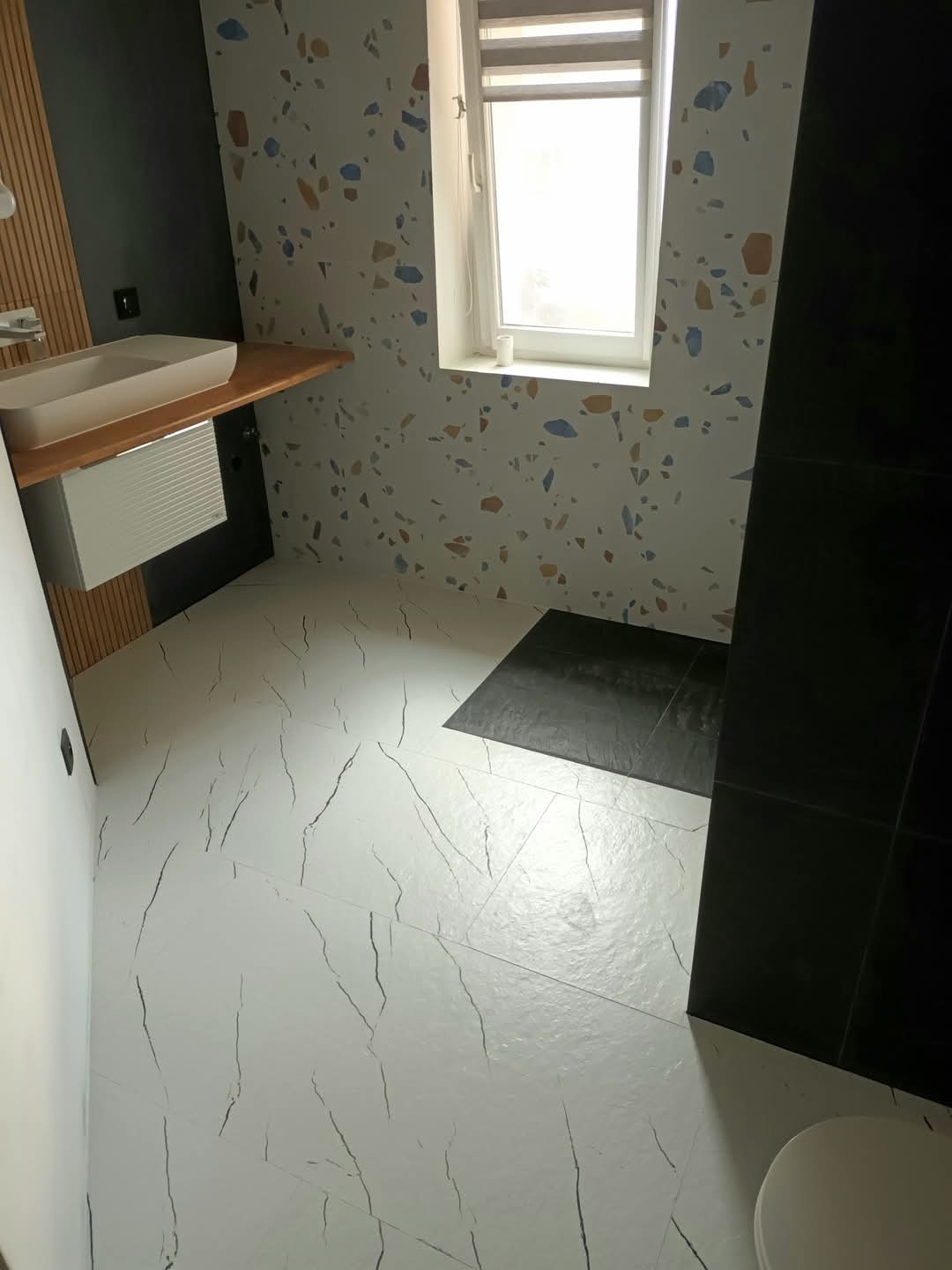 Nowoczesna łazienka po renowacji: białe płytki z czarnymi żyłkami na podłodze, ściana z wzorem lastryko, drewniany blat z umywalką i czarne akcenty.