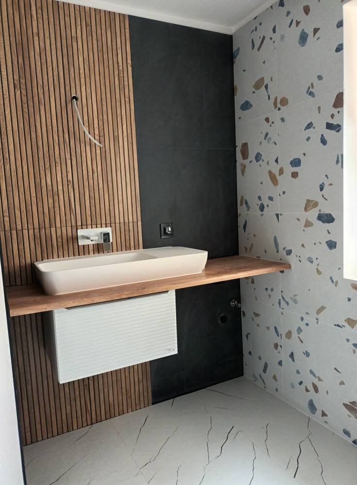 Nowoczesna łazienka z umywalką nablatową, drewnianą boazerią, czarnymi płytkami i mozaiką. Minimalistyczny design, jasne wnętrze, marmurowa podłoga.