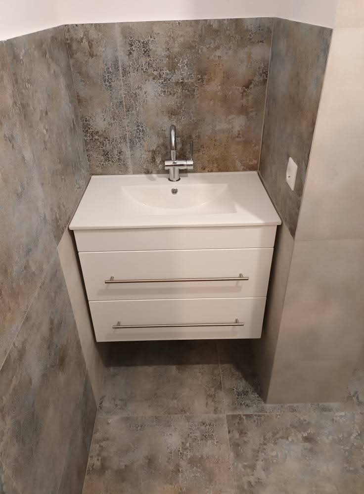 Nowoczesna łazienka: biała szafka pod umywalką z chromowaną baterią, tło z płytek imitujących kamień w odcieniach szarości i beżu. Minimalistyczny design.