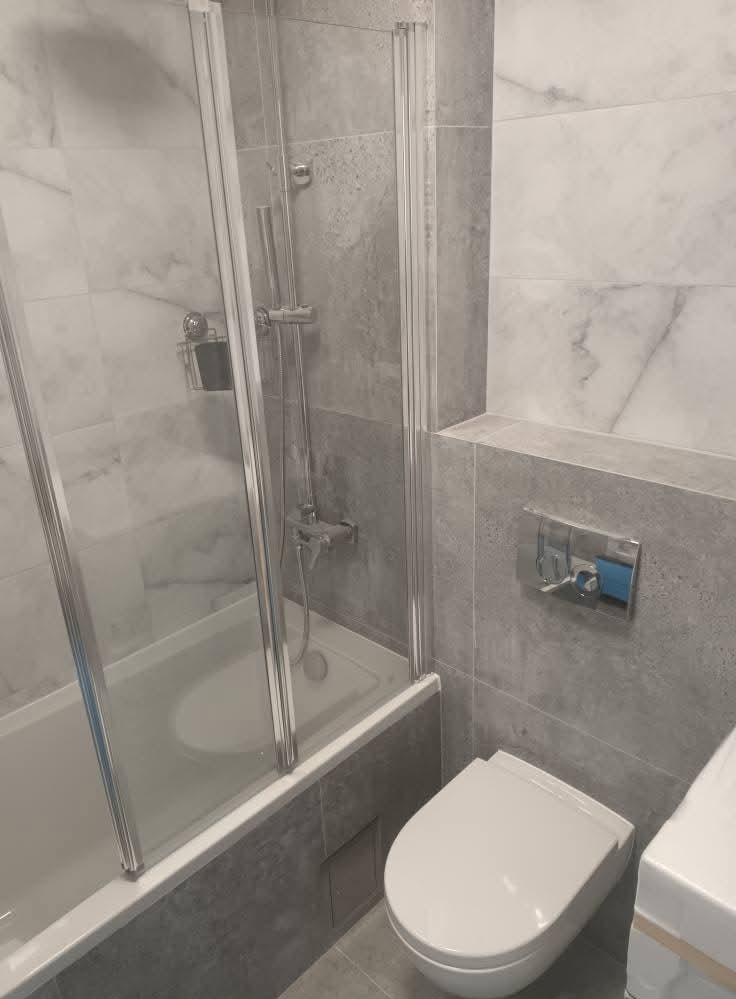 Szara łazienka po remoncie: kabina nawannowa ze srebrnymi profilami, toaleta podwieszana i marmurowe płytki. Minimalistyczny design z akcentem na funkcjonalność.