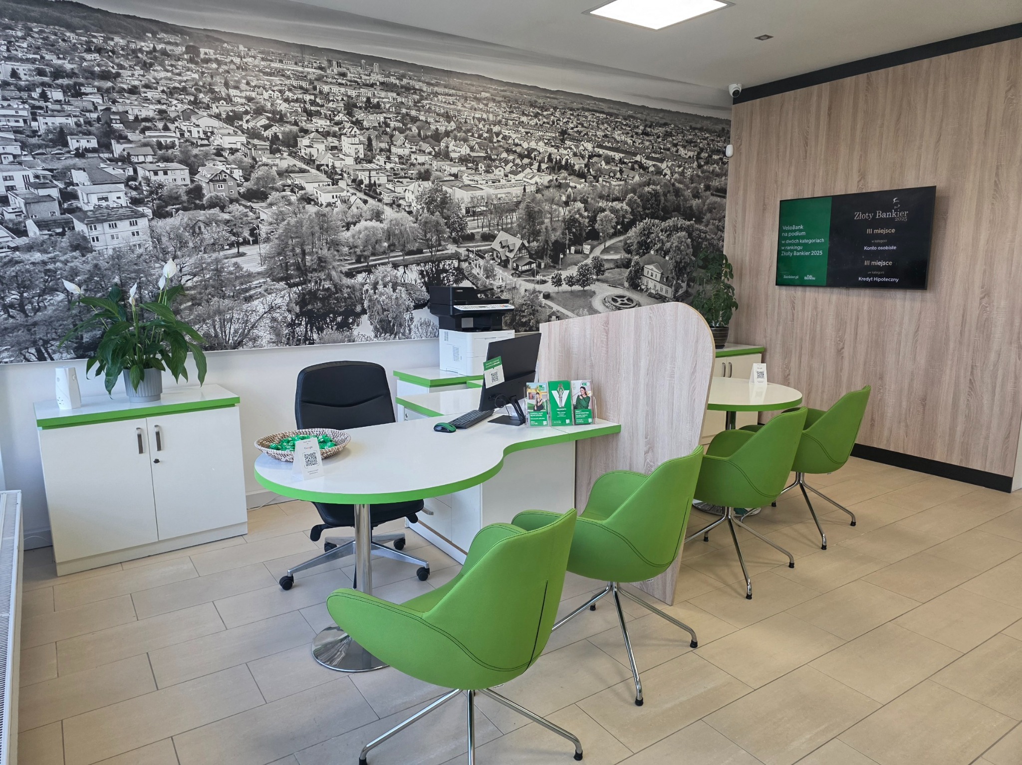 Nowoczesne biuro doradcy finansowego z zielonymi krzesłami i fototapetą miasta. Na ścianie tablica z nagrodą 'Złoty Bankier 2025' za konto osobiste i kredyt hipoteczny.