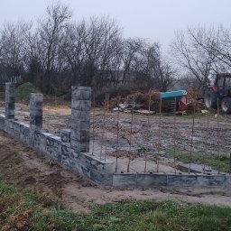 Dominik Chmielewski - Wznoszenie ogrodzenia z szarej kostki betonowej. Widoczne słupki i podmurówka w trakcie budowy. W tle traktor i teren budowy w pochmurny dzień.