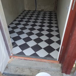 Dominik Chmielewski - Ułożona podłoga z czarno-białych płytek w szachownicę w małym pomieszczeniu. Widoczny fragment drzwi i białe wiadro.