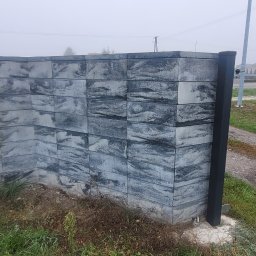 Dominik Chmielewski - Nowoczesne ogrodzenie betonowe z elementami metalowymi w szarym odcieniu, widoczne z bliska w pochmurny dzień. Charakterystyczna faktura bloczków imitująca kamień.