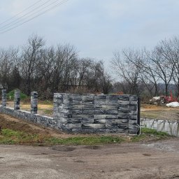 Dominik Chmielewski - Fragment ogrodzenia betonowego z szarej kostki, widok z perspektywy ulicy w pochmurny dzień. Część ogrodzenia w trakcie budowy, widoczne słupki i fragment muru.