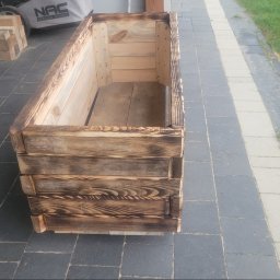 Dominik Chmielewski - Drewniana skrzynia na kwiaty z opalonymi brzegami, ustawiona na szarym bruku. W tle trawnik i kosiarka. Widoczne detale konstrukcji i faktura drewna.